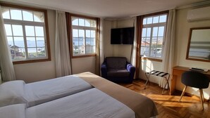Habitación de hotel con dos camas, televisión, una silla, escritorio y ventanas amplias con vista al mar.