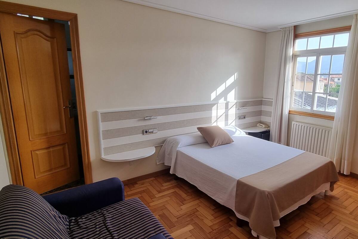 A hotel room with a bed, a bedside table, a chair, a desk, and a window with a view.