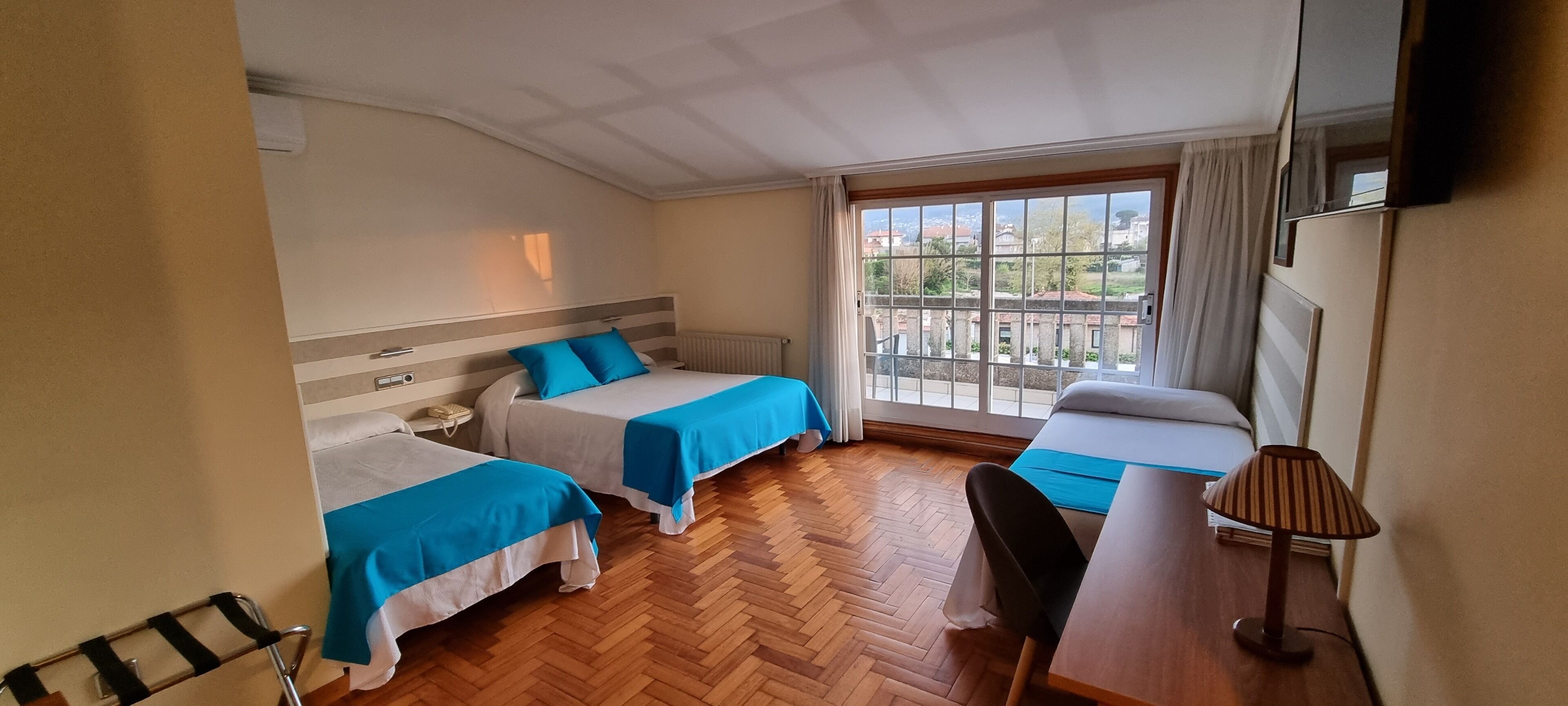 A hotel room with two beds, a wooden floor, a desk, a chair, a lamp, and a window with a view of buildings.