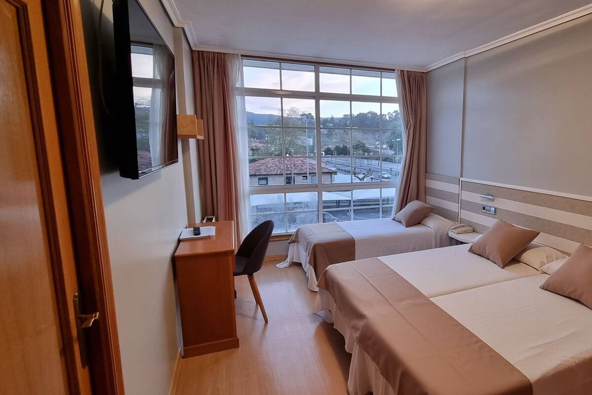 A hotel room with two beds, a desk, and a large window offering a view of the cityscape.