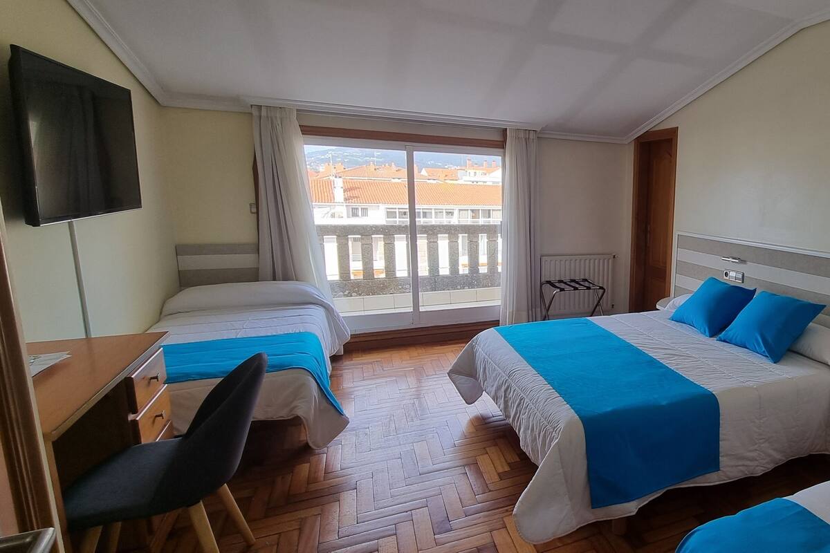 A hotel room with two beds, a desk, a chair, a TV, and a view of buildings through a large window.