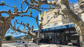 Un patio con un edificio que tiene ventanas amplias y fachada de piedra. Hay sillas y mesas al aire libre, y un árbol con ramas desnudas.