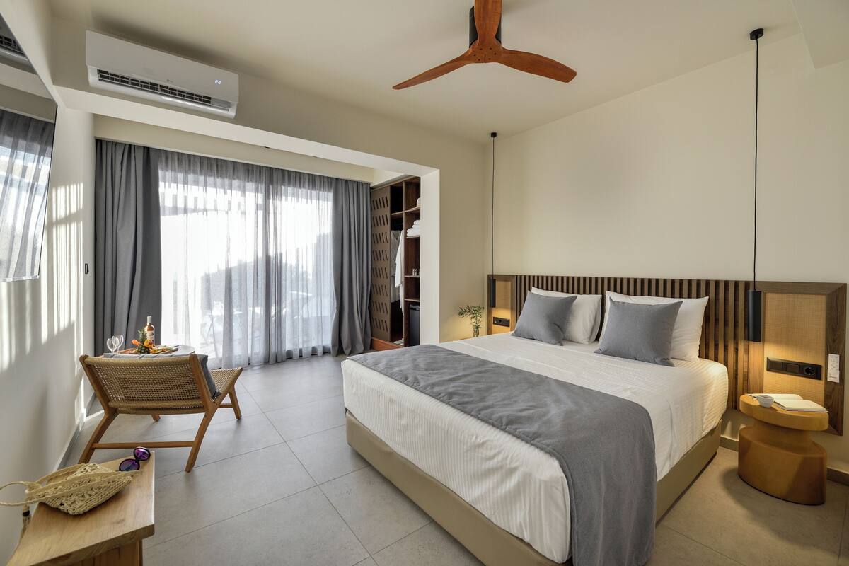 Een moderne hotelkamer met een groot bed, een houten plafondventilator, een zithoek met een tafel en stoelen, en een inbouw kast.