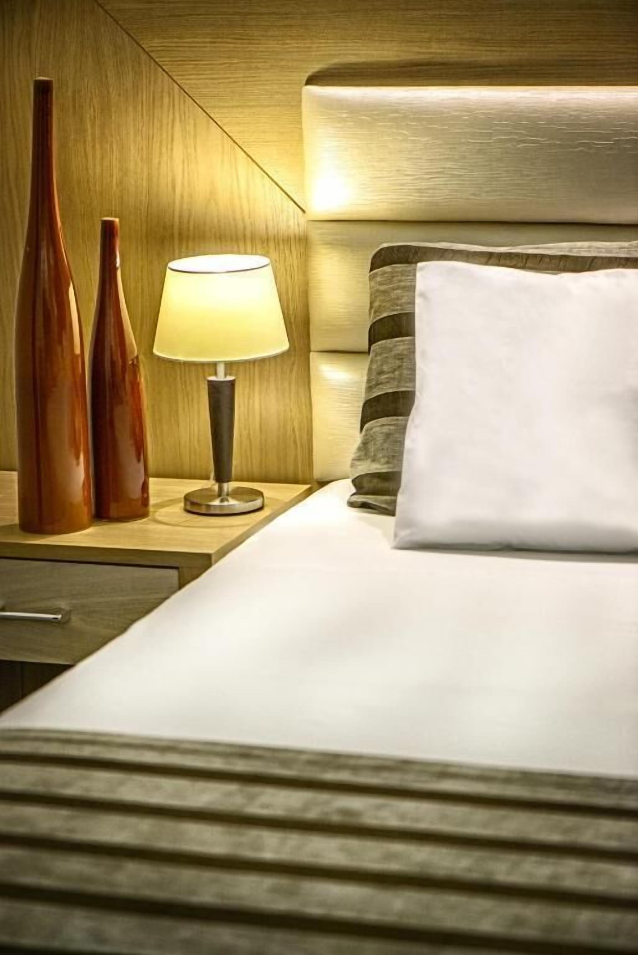 A hotel room with a wooden headboard, a bedside lamp, two decorative vases, and a white pillow.