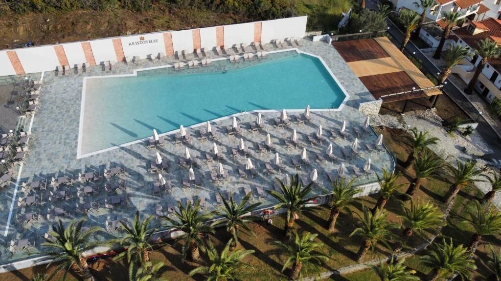 A pool area with lounge chairs and umbrellas, surrounded by palm trees and a building labeled "ARISTOTLE."