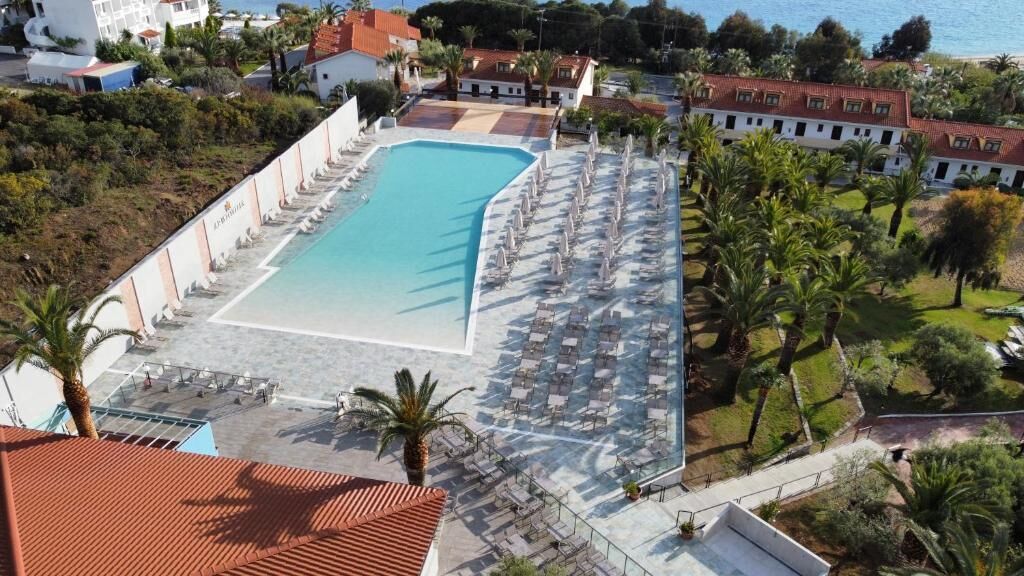 Una piscina rodeada de sillas de descanso y sombrillas, con vista al mar y edificios al fondo.