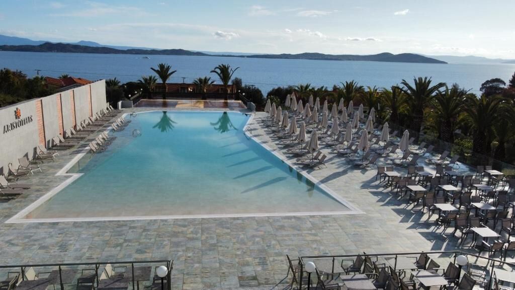 Una piscina con sillones de descanso y sombrillas, vista al mar, y un edificio con un logotipo.