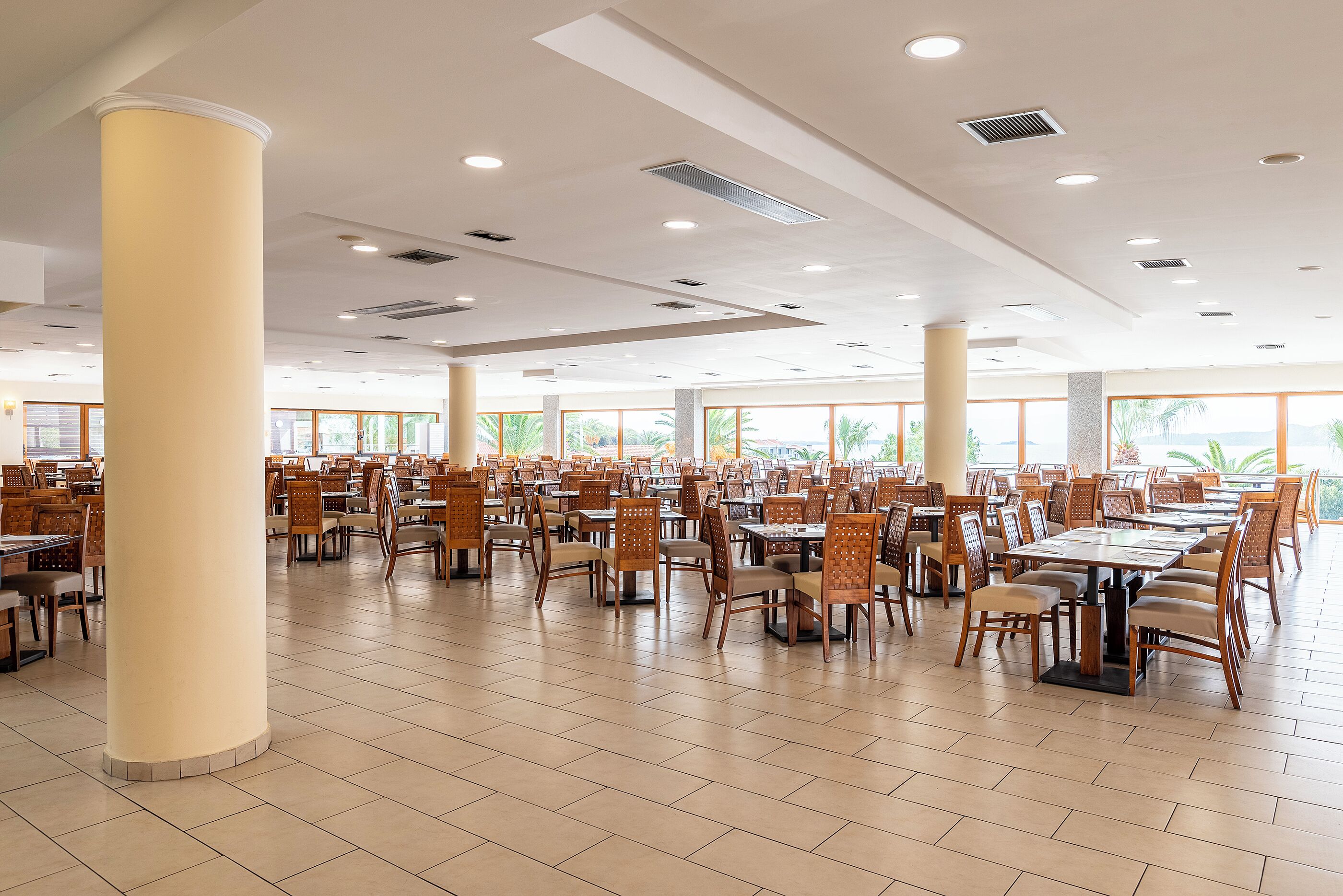 A spacious dining area with multiple tables and chairs, large pillars, and a high ceiling with recessed lighting.