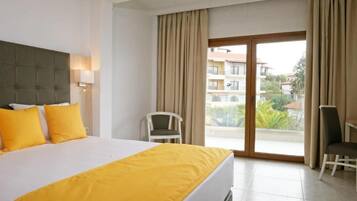 A hotel room with a bed, a chair, a desk, and a balcony with a view of buildings and greenery.