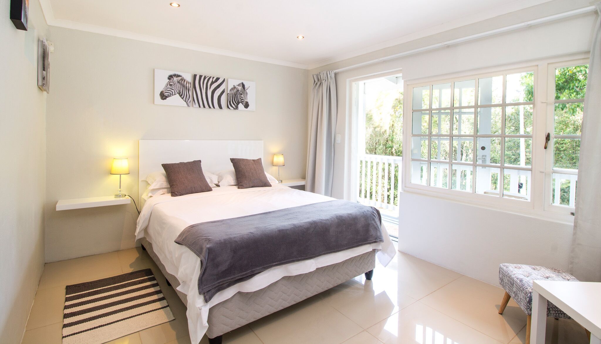 A bedroom with a bed, bedside lamps, a zebra artwork, a window with curtains, and a small table.