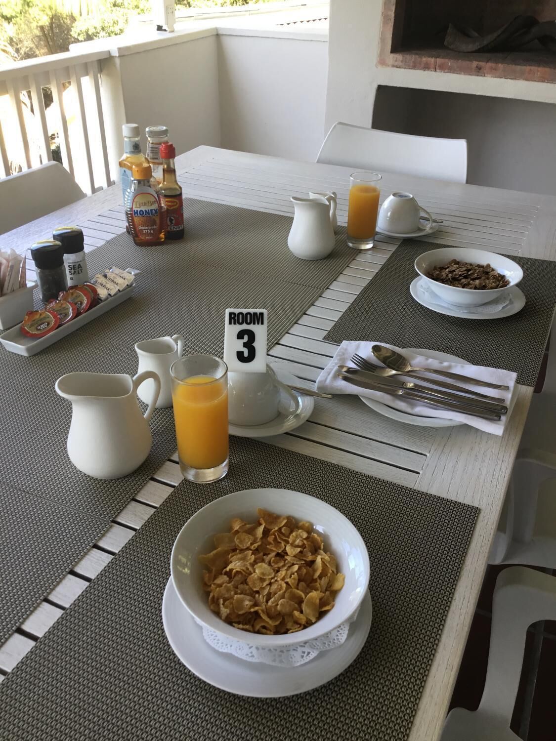 Mesa de desayuno en una habitación con un letrero que indica 'ROOM 3', con cereales, jugo, miel y condimentos.