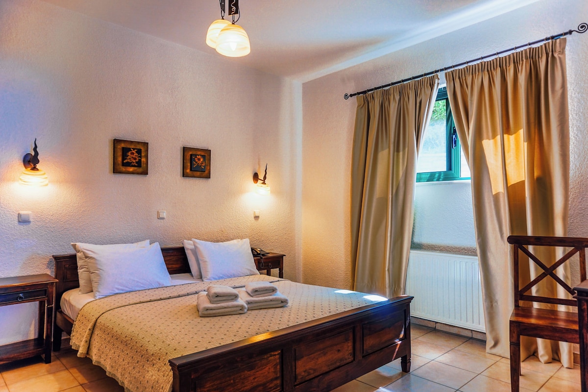 A neatly made bed with a patterned bedspread and pillows, a wooden nightstand with a lamp, and a window with curtains.