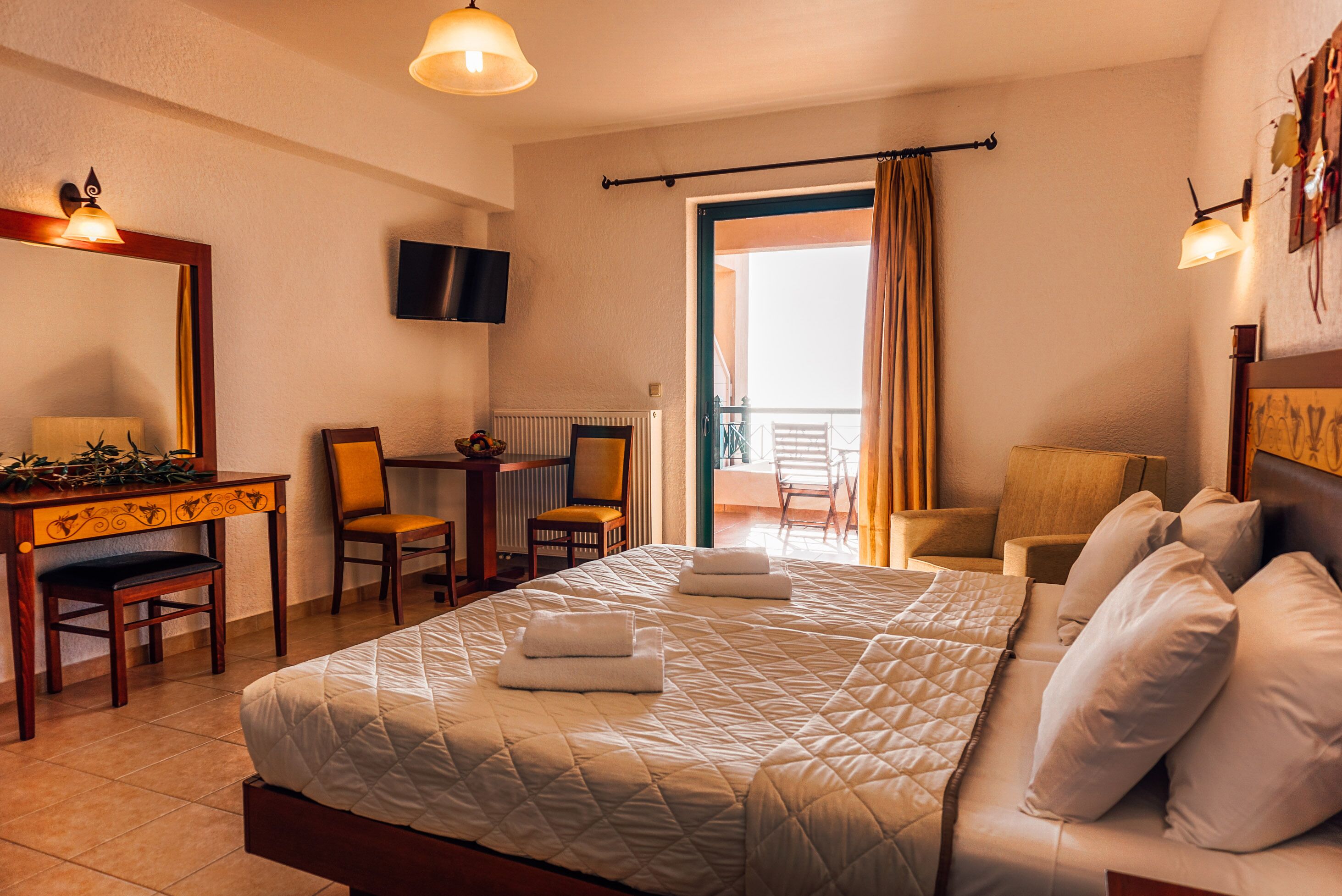 A hotel room with a large bed, a desk with a chair, a TV mounted on the wall, and a balcony with curtains.