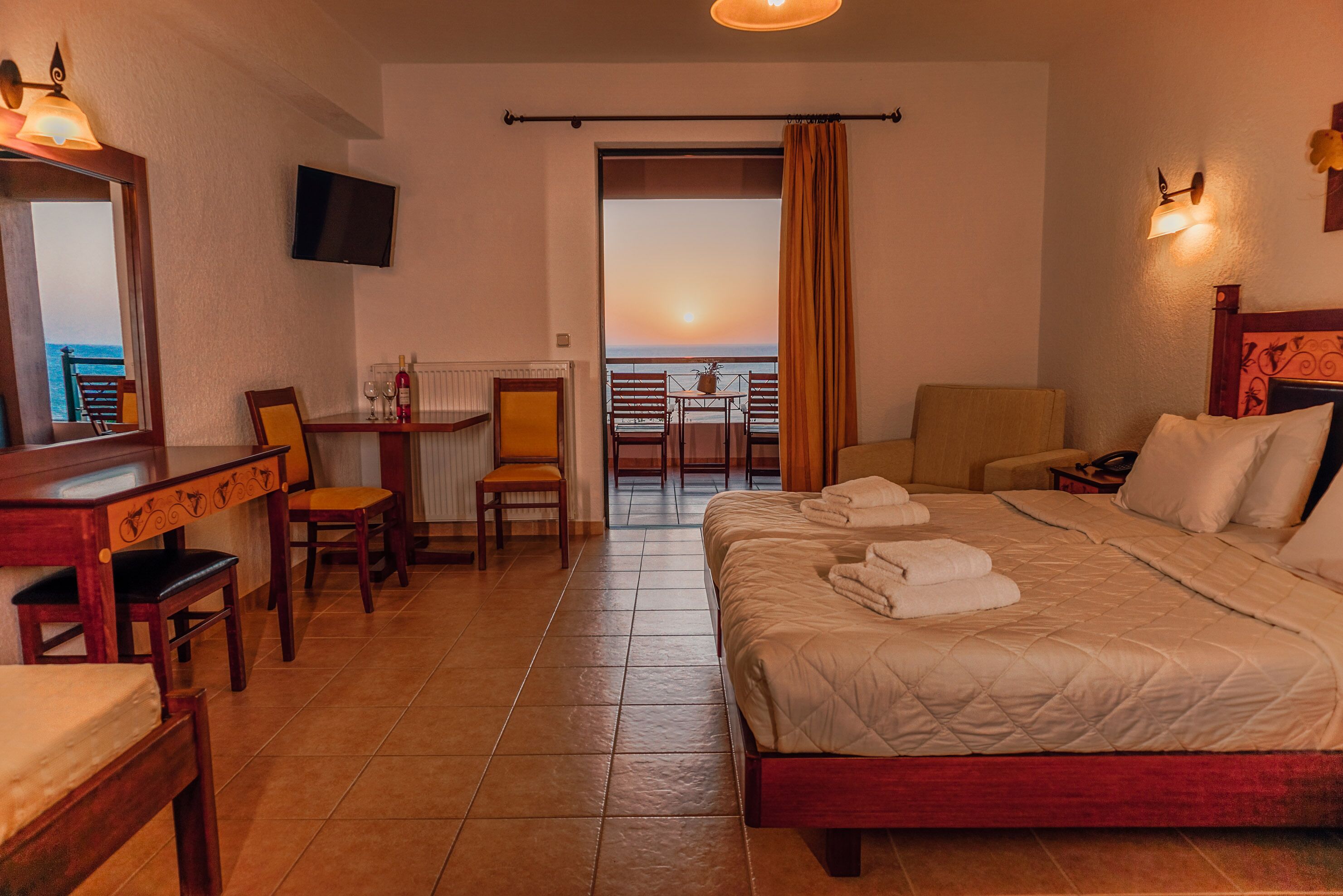A hotel room with a bed, a desk, a chair, a TV, and a balcony with a view of the sunset.