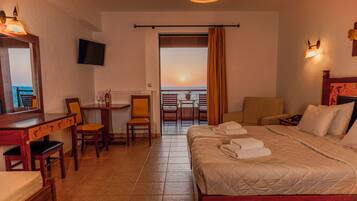A hotel room with a bed, a desk, a chair, a TV, and a balcony with a view of the sunset.