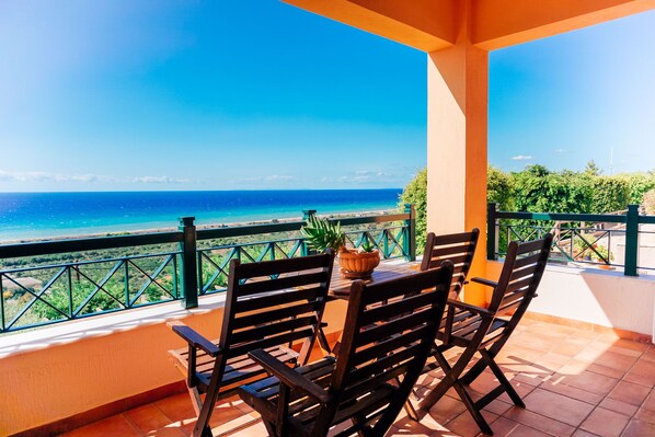 Un balcón con mesa y sillas, con vista al mar azul y la costa lejana.