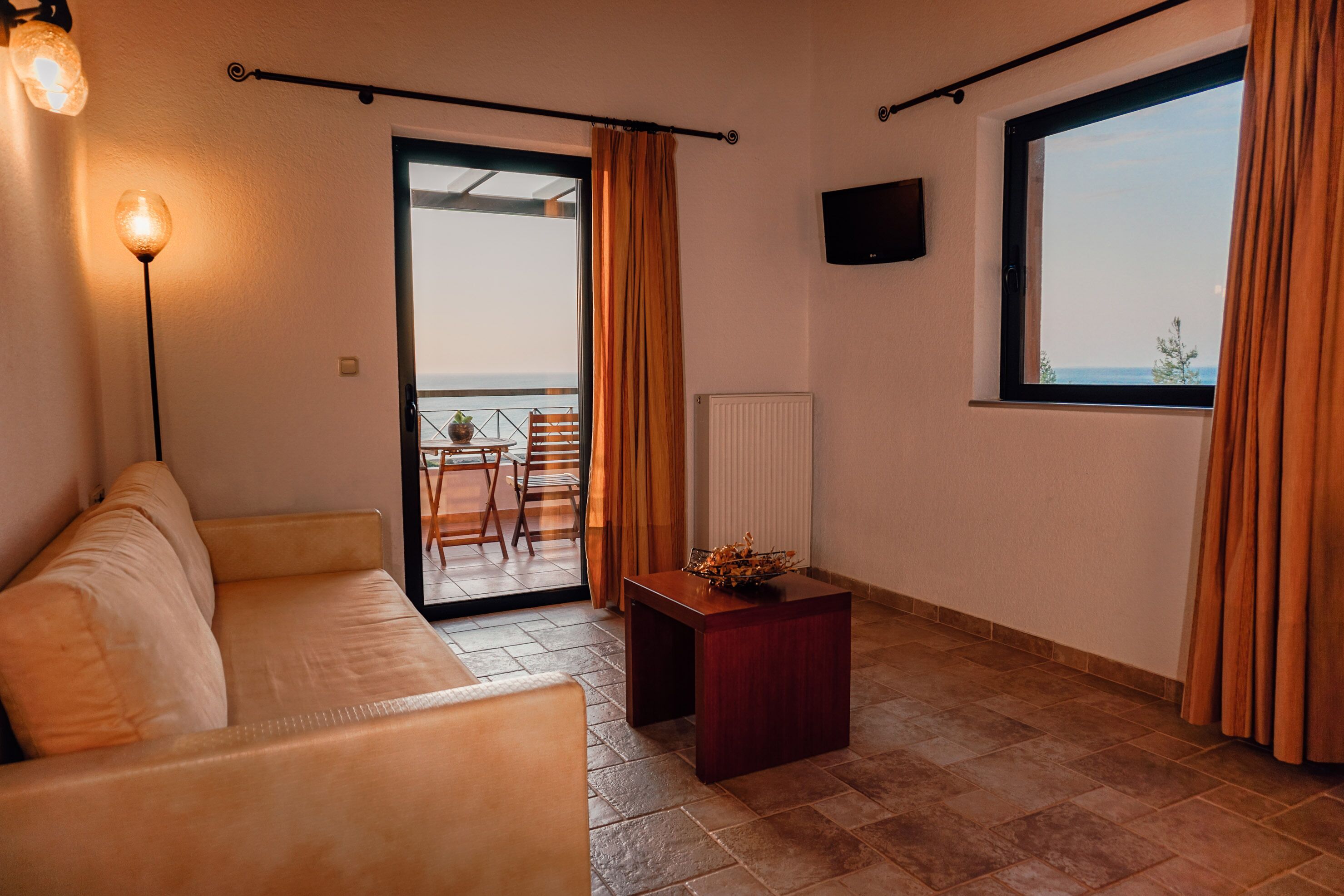 A living room with a sofa, a small table, a TV mounted on the wall, and a view of the outdoors through a sliding door.