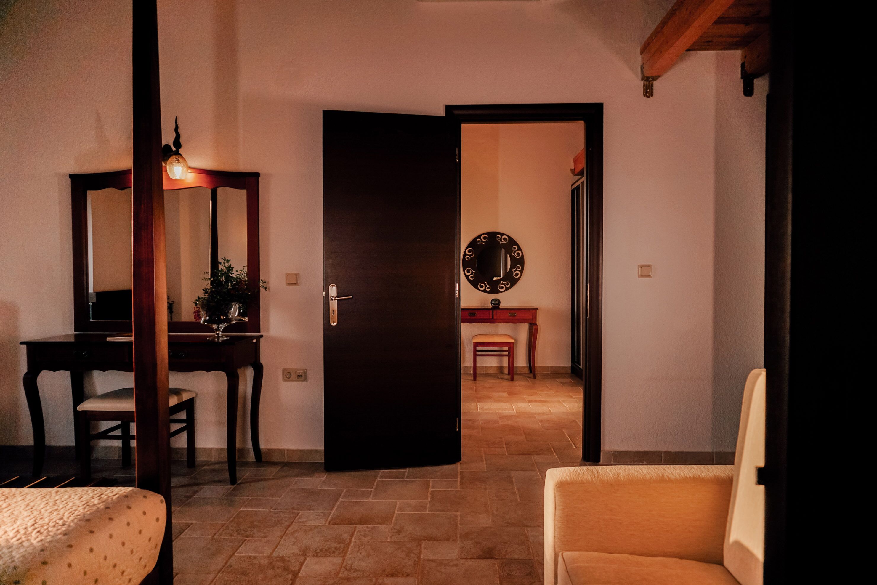 A room with a desk, chair, and a bed. There is a door leading to another room with a table and a chair.