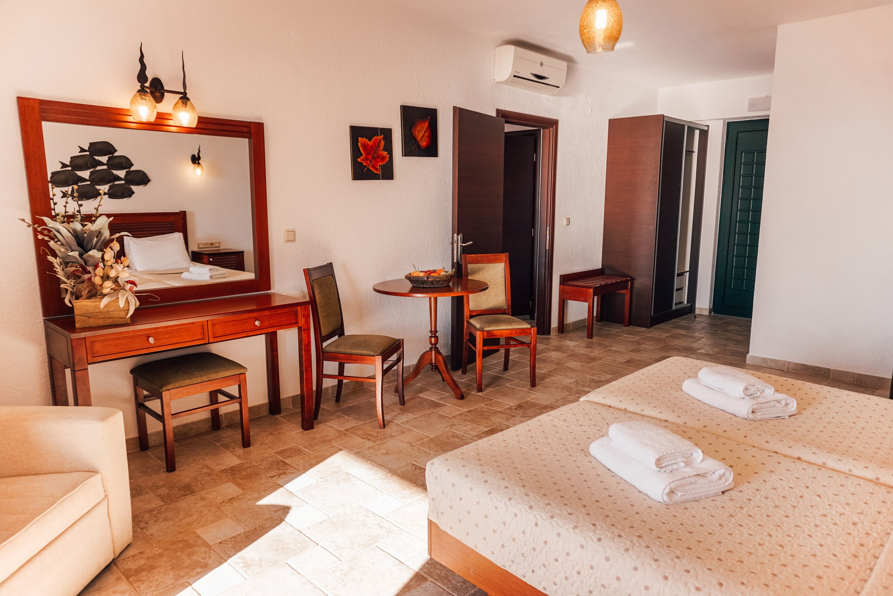 A hotel room with a bed, a desk, a chair, a small table with a bowl of fruit, a mirror, and a wall-mounted air conditioner.