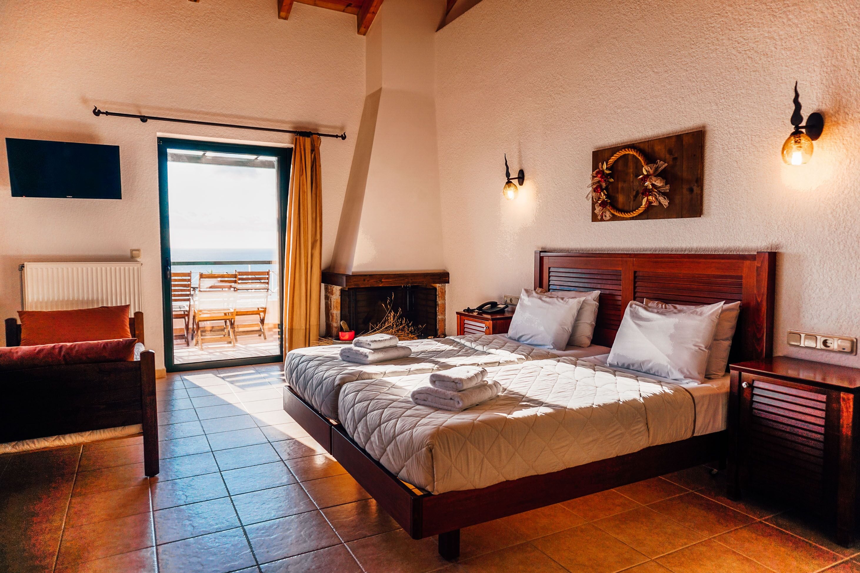 A bedroom with a large bed, a wooden headboard, a fireplace, a TV mounted on the wall, and a balcony with a view.