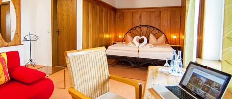 Habitación de hotel con cabecera de madera, cama con almohadas en forma de corazón, sofá rojo, silla de mimbre y un ordenador portátil sobre una mesa de madera.