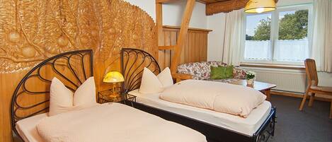 A room with two beds, a wooden headboard with a carved leaf design, a window with curtains, and a table with a lamp.