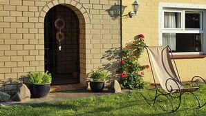 Une maison avec une façade en briques, une porte cintrée de couleur sombre et une fenêtre à cadre blanc. On peut voir des plantes en pot et un banc en métal à l’extérieur.