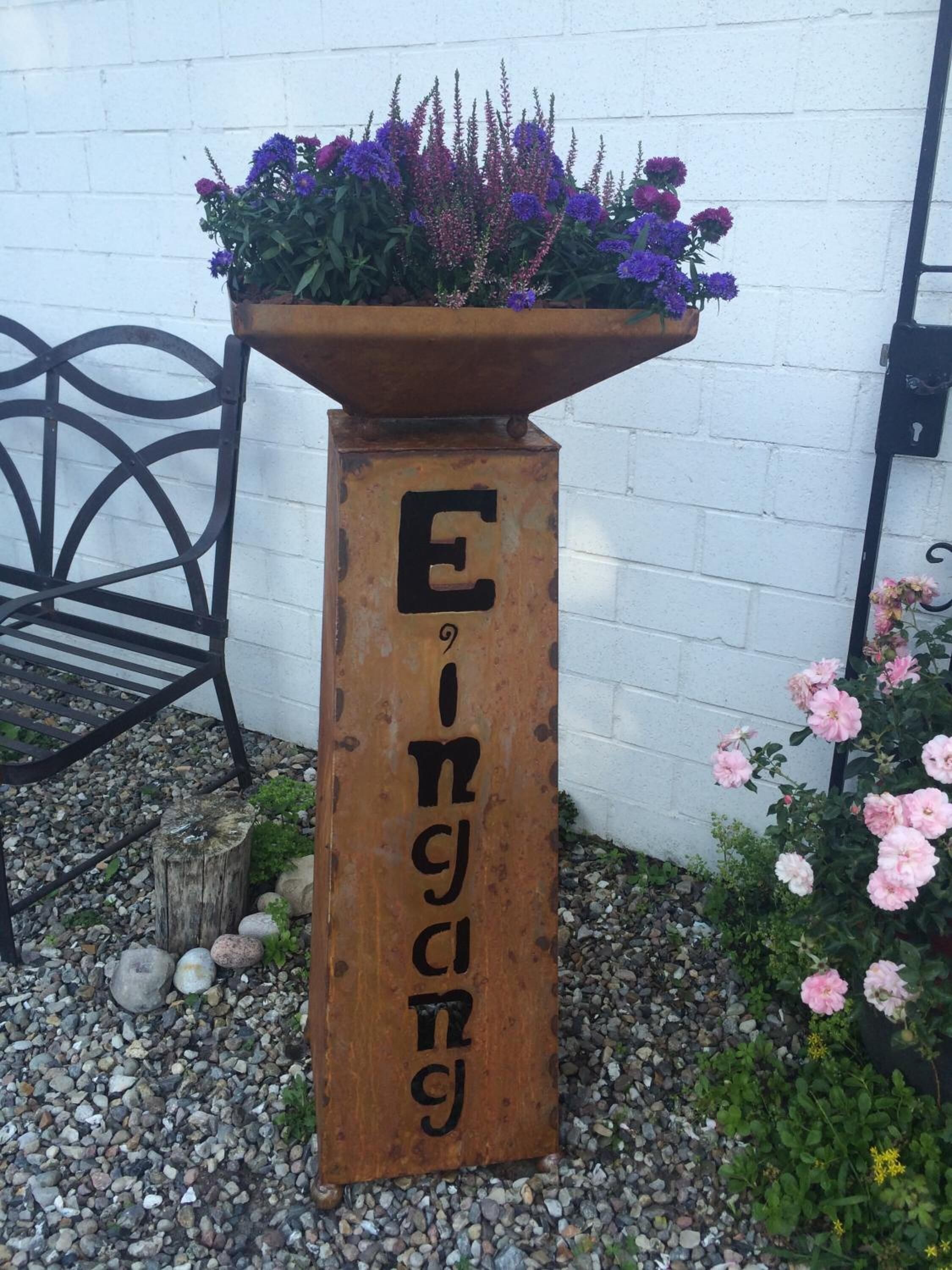 A wooden planter with flowers and the text "Eingang" against a white wall.