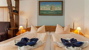 Une chambre d’hôtel avec deux lits, chacun agrémenté d’une serviette et d’un oreiller. Un tableau représentant un voilier est accroché au mur, et une échelle en bois est appuyée contre le mur.