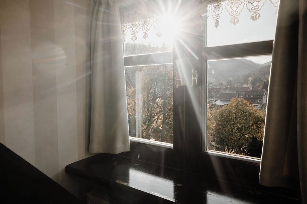 A room with a piano near a window with curtains, a view of a town and mountains outside.
