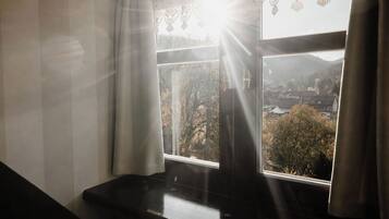A room with a piano near a window with curtains, a view of a town and mountains outside.