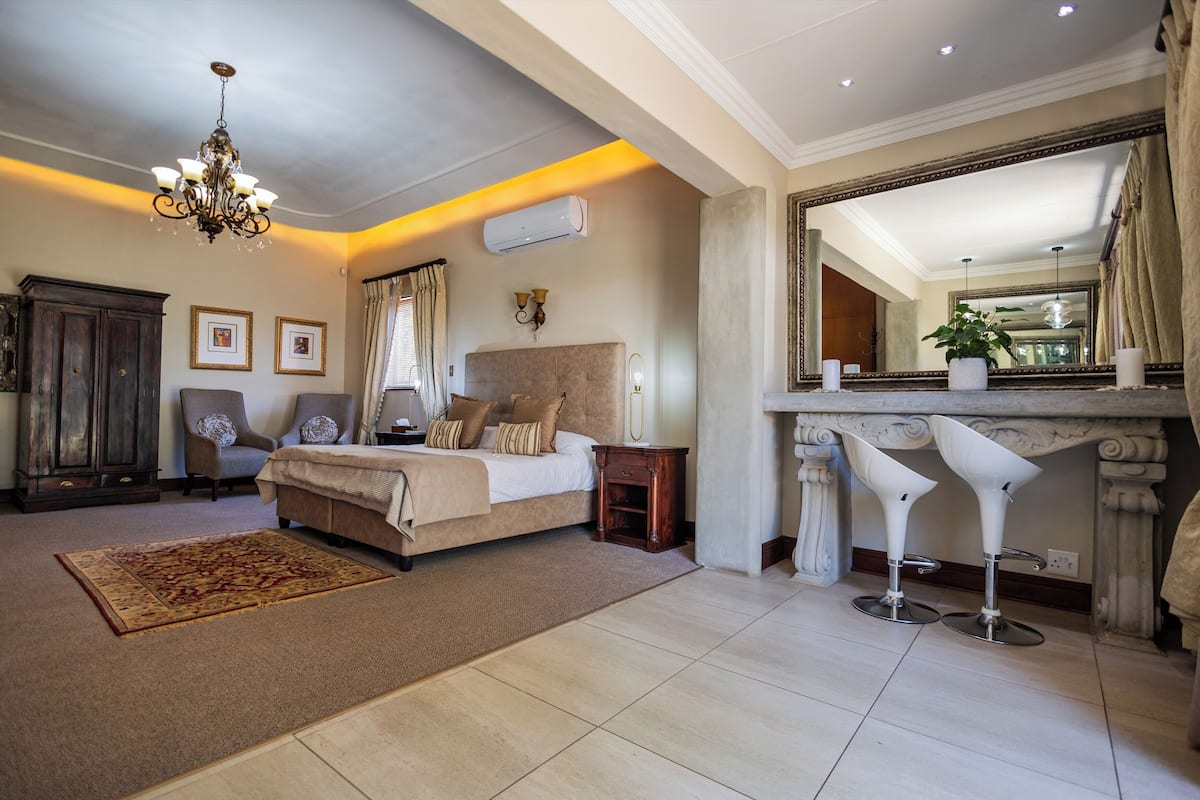 A bedroom with a large bed, a chandelier, a mirror, and two bar stools.