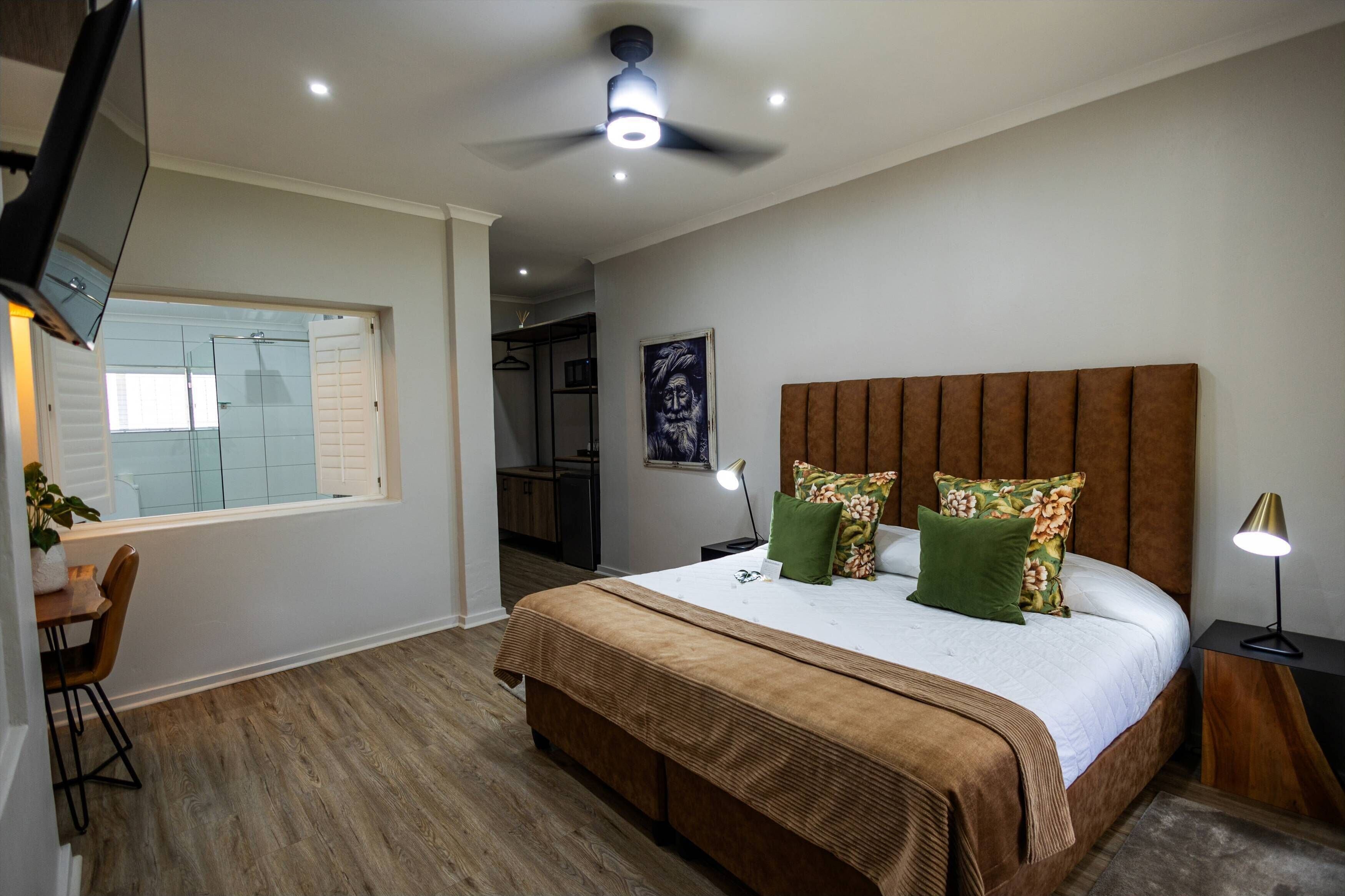 Ein Schlafzimmer mit einem großen Bett, einem Deckenventilator, einem an der Wand montierten Fernseher und einem Fenster mit Blick auf ein Badezimmer.