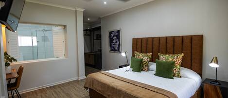 Ein Schlafzimmer mit einem großen Bett, einem Deckenventilator, einem an der Wand montierten Fernseher und einem Fenster mit Blick auf ein Badezimmer.