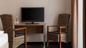Habitación de hotel con una televisión sobre un escritorio de madera, dos sillas de mimbre, una botella de agua y una cama.
