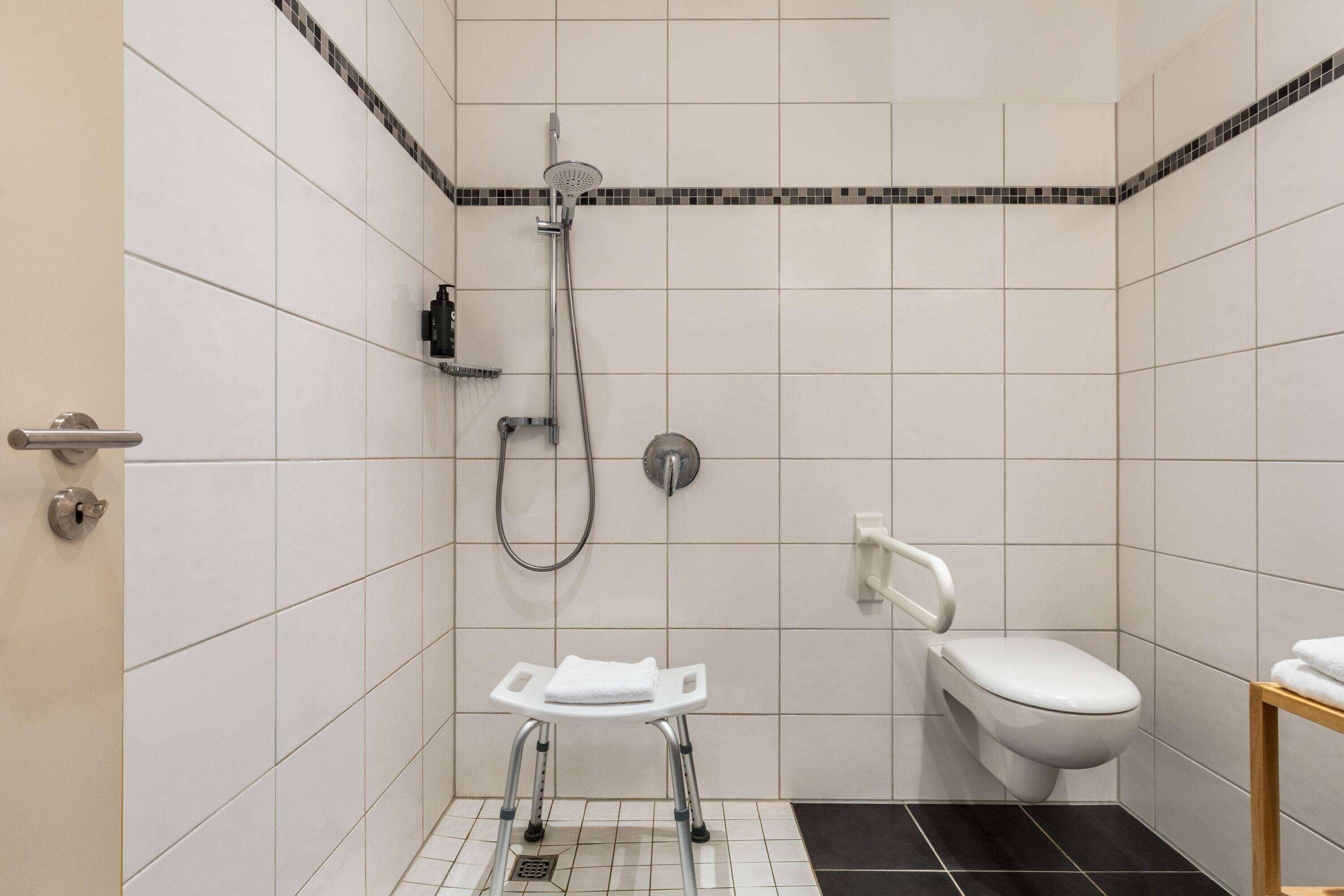 A small, modern bathroom with white tiles, a shower, a toilet, and a grab bar.