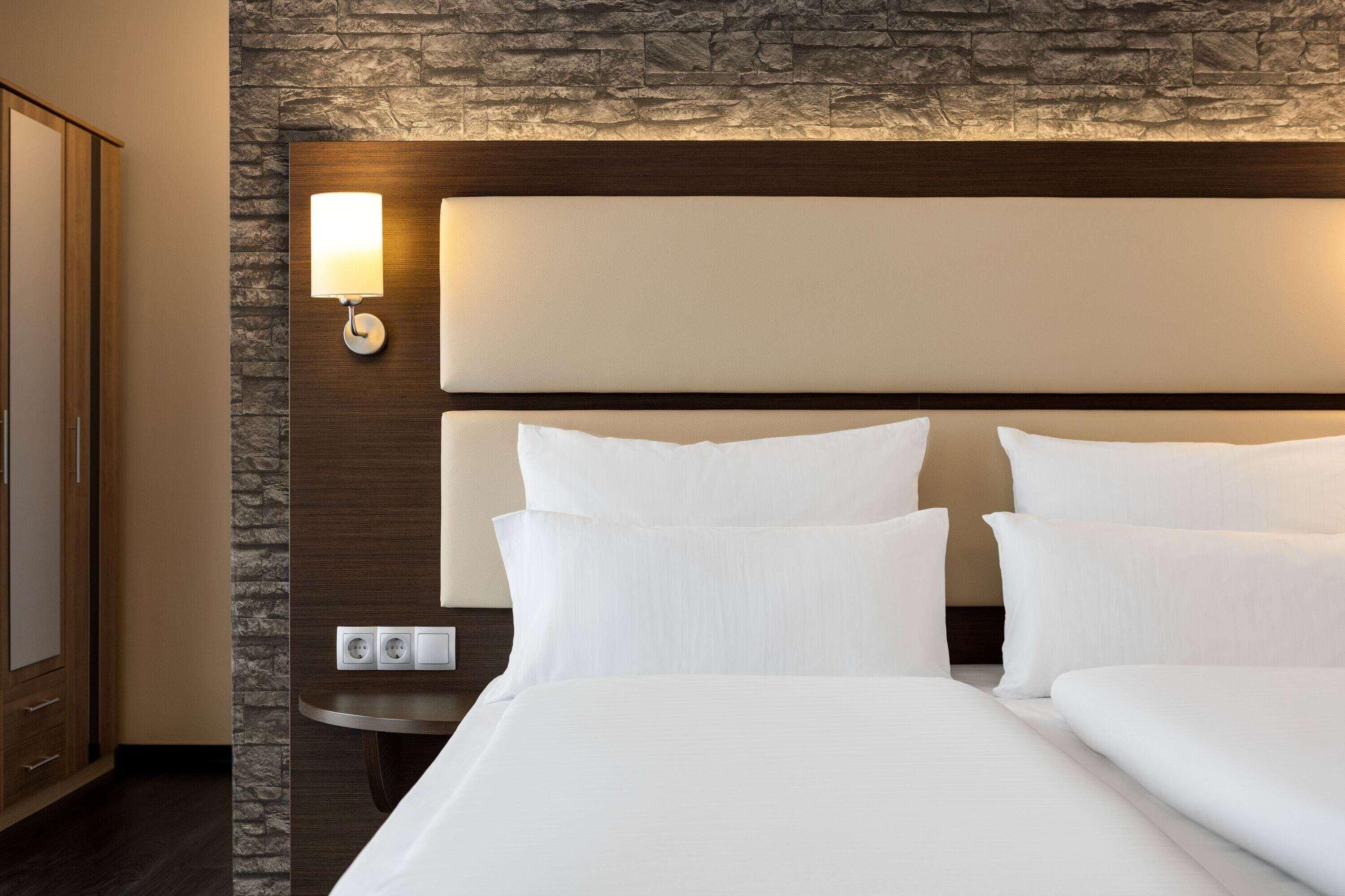 Quarto de hotel com uma cama grande, cabeceira com luminária, parede de destaque em pedra e pequena mesa de cabeceira.