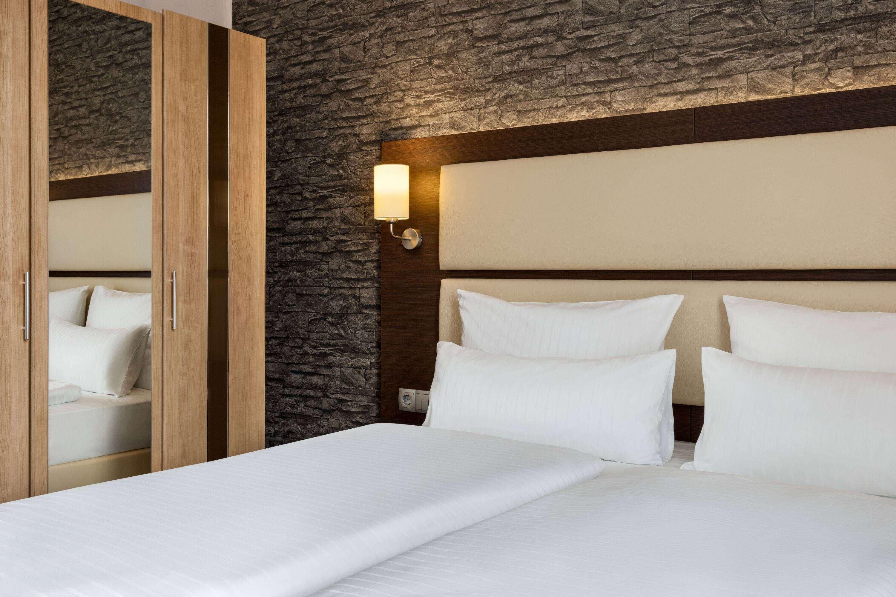 A hotel room with a stone wall, a wooden wardrobe, a bed with white linens, and a wall-mounted light fixture.