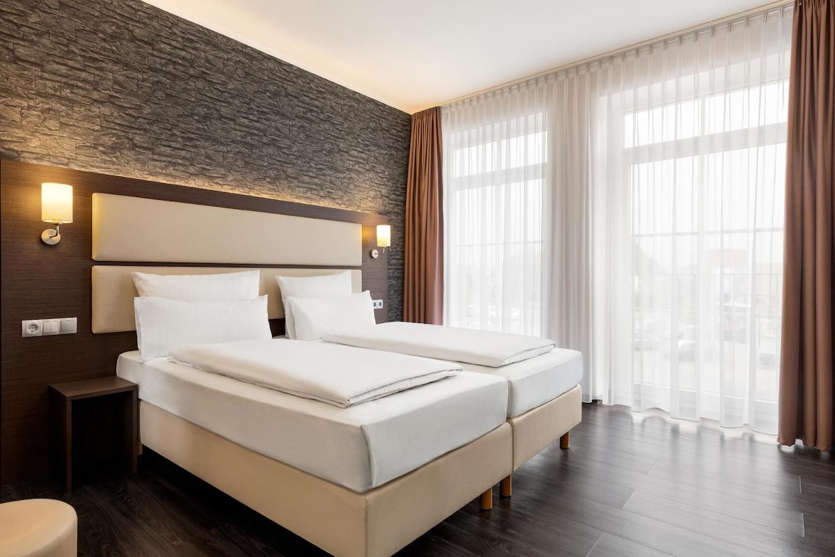 A hotel room with a double bed, a nightstand, a wall-mounted lamp, and a large window with curtains.