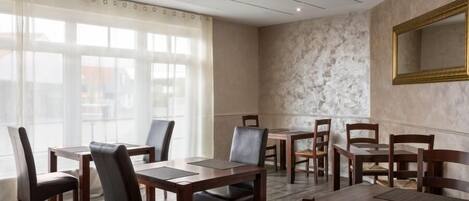 A dining area with multiple tables and chairs, large windows with curtains, and a marble wall with a framed mirror.