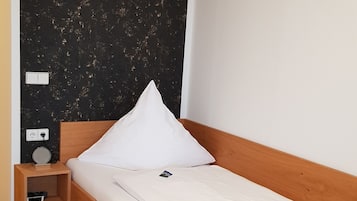 A single bed with a wooden frame and white bedding, a bedside table with a lamp, and a power outlet.