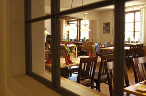En interiörbild av en restaurang med träbord och stolar, hängande lampor och en utsikt genom ett fönster.