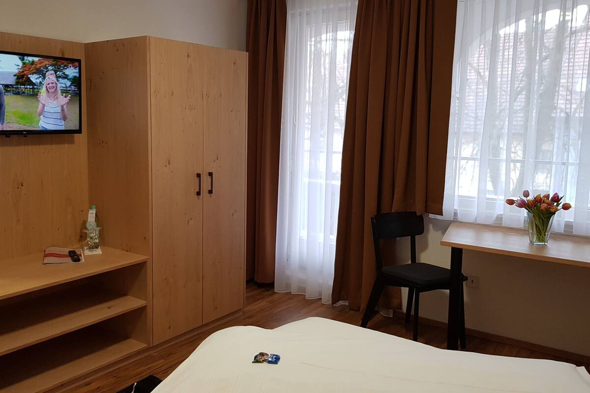 Une chambre d’hôtel équipée d’une télévision, d’une armoire en bois, d’un bureau avec une chaise, d’un lit et d’une fenêtre avec des rideaux.