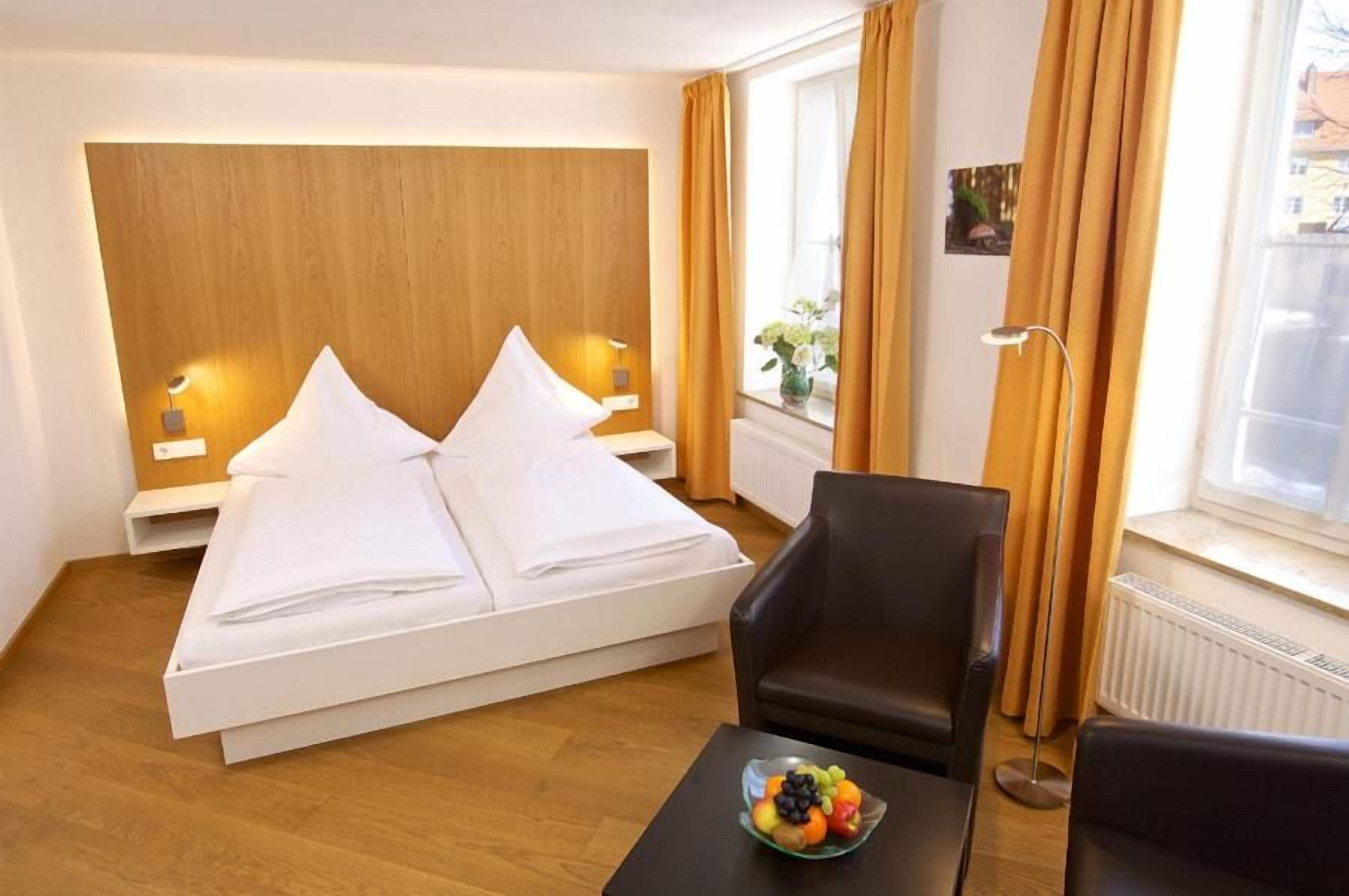 A hotel room with a bed, a chair, a table with fruit, and a window with curtains.