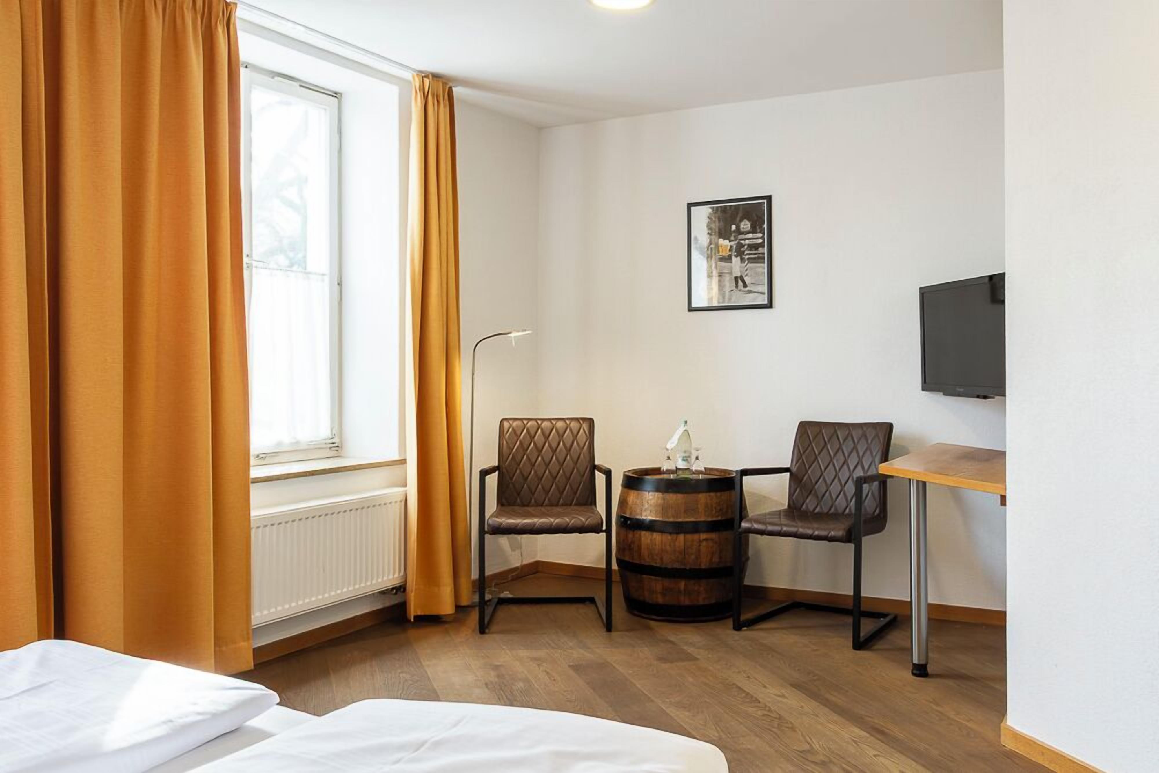 A hotel room with a bed, two chairs, a small table, a barrel, a radiator, a window with curtains, a picture on the wall, and a TV on the wall.
