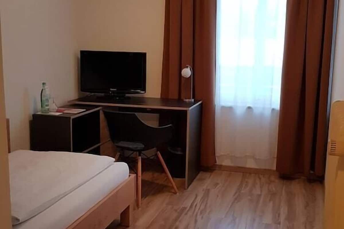 Une chambre d’hôtel comprenant un lit, un bureau avec une chaise, une télévision et une fenêtre avec des rideaux.