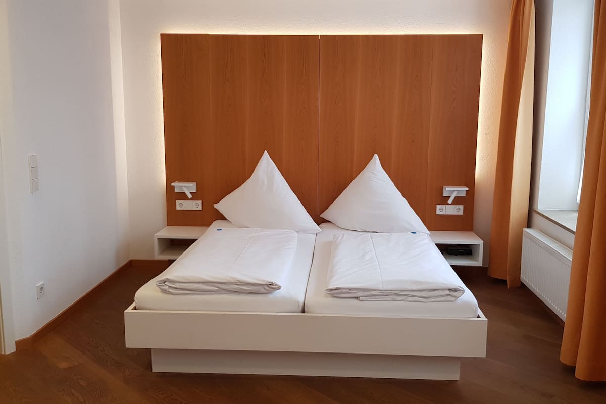 Un lit double avec une literie blanche, une tête de lit en bois, des tables de chevet avec des lampes et une fenêtre avec des rideaux.