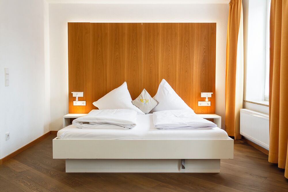 A modern hotel room with a wooden headboard, white bedding, two bedside tables, and a window with curtains.