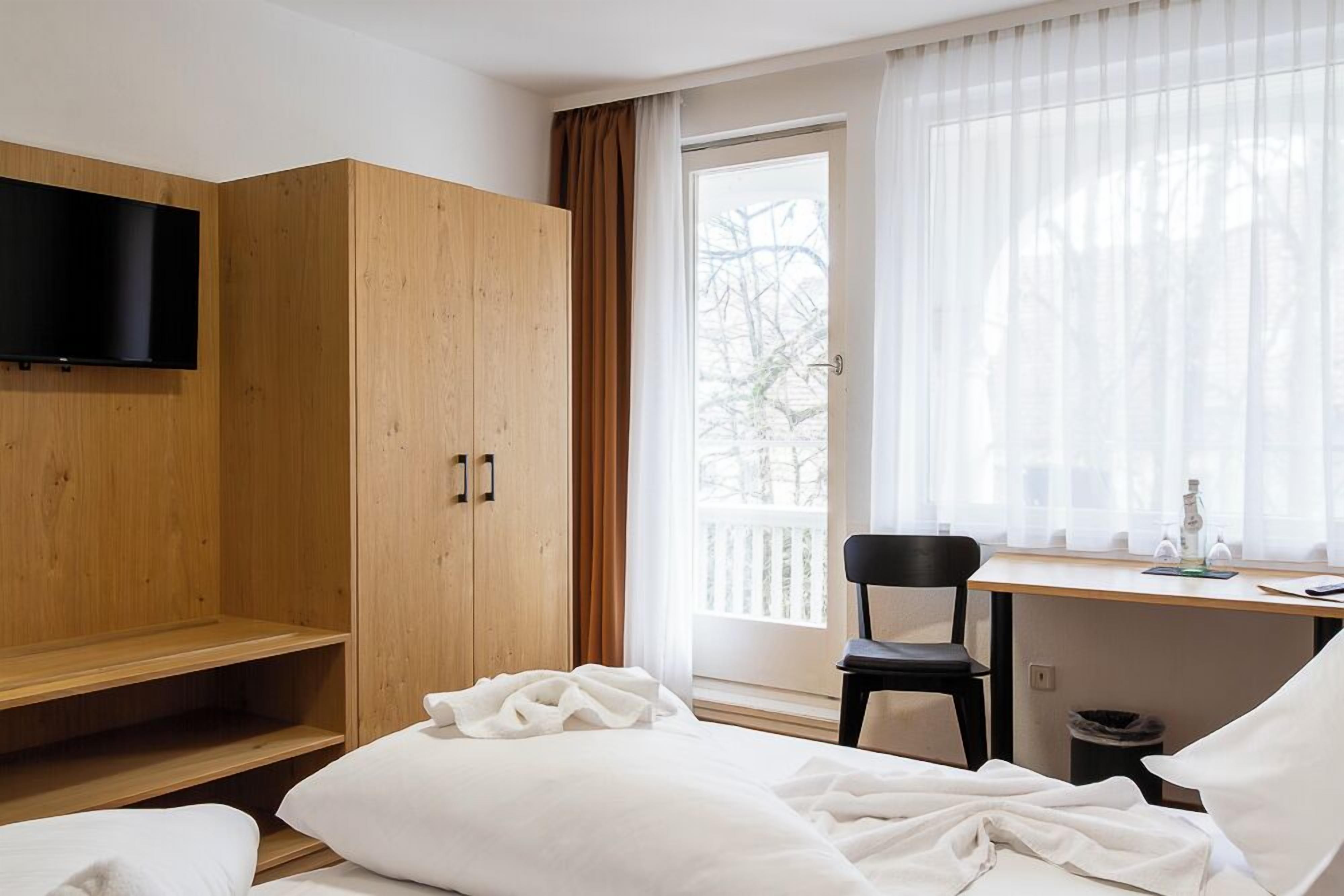 A hotel room with a bed, a wooden wardrobe, a desk with a chair, a television, and a window with curtains.