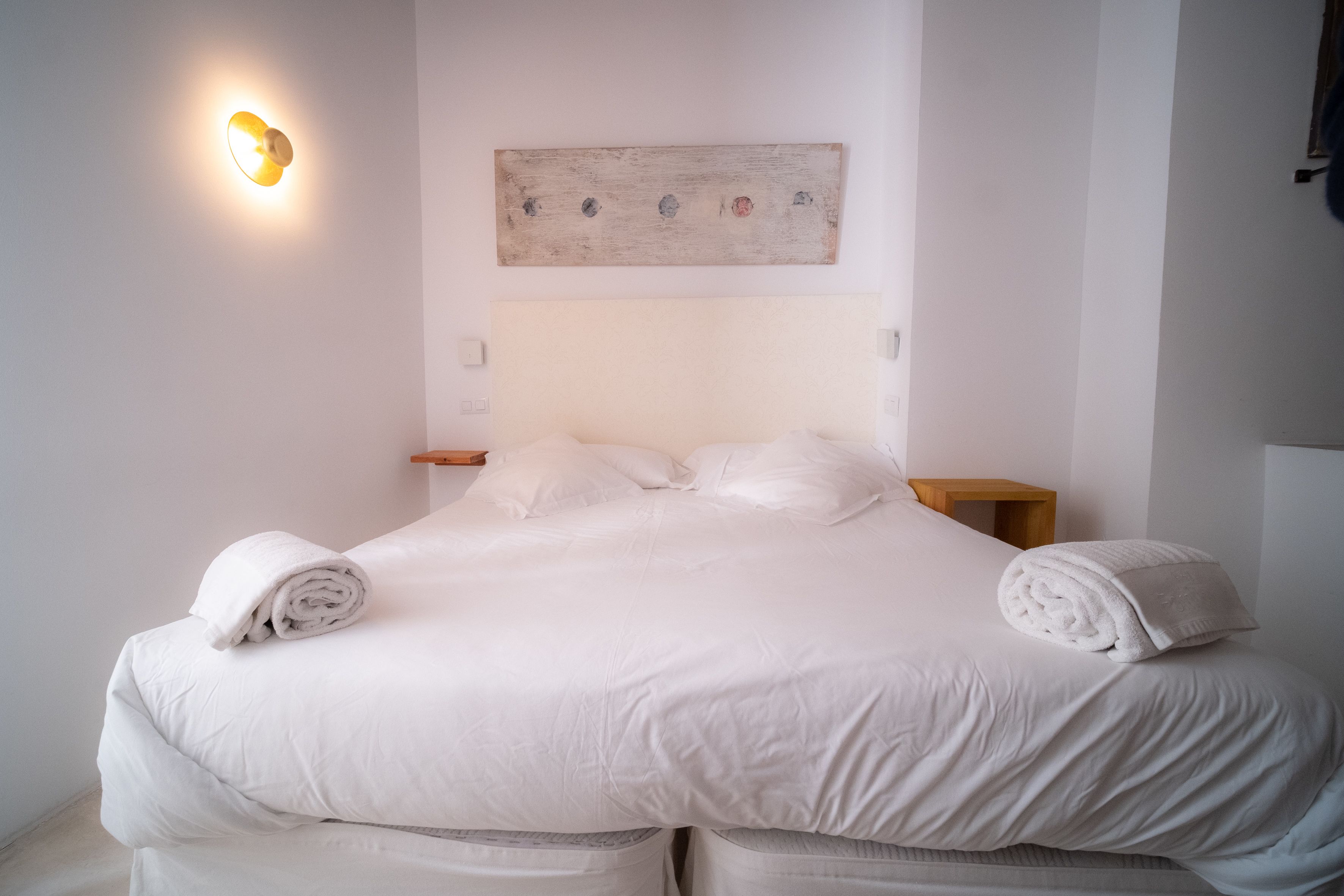 Un lit bien fait, avec du linge de lit blanc et deux serviettes enroulées de chaque côté, une lampe fixée au mur et un tableau simple au-dessus du lit.